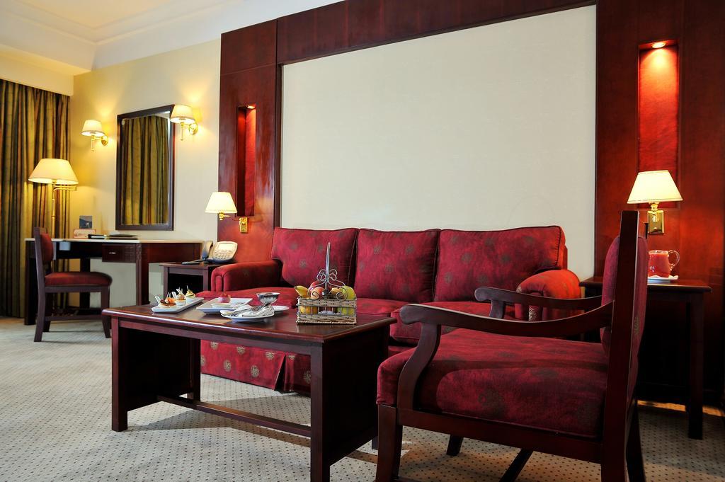 Safir Hotel Cairo Giza Room photo The photo shows a well-decorated hotel room or sitting area. In the center, there is a red couch with matching armchairs around it. A wooden coffee table is placed in front of the couch, and it holds some decorative items and snacks. The walls are ad