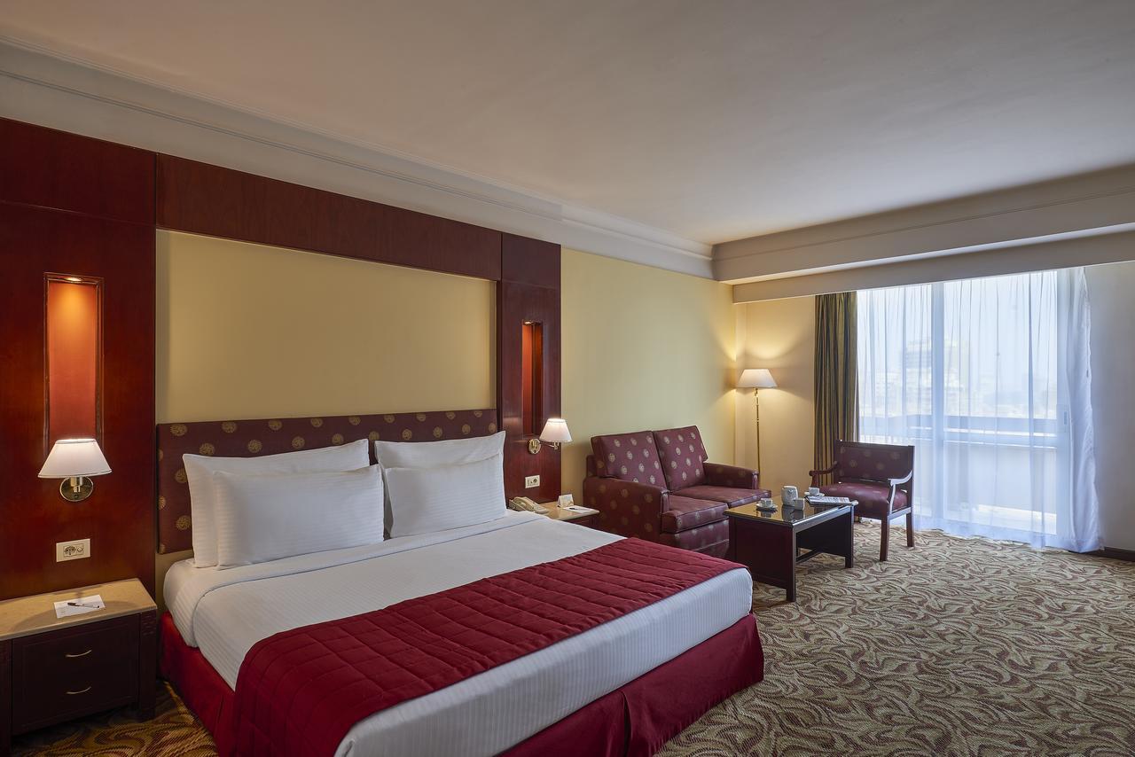 Safir Hotel Cairo Giza Exterior photo The image showcases a hotel room featuring a large bed with white linens and a red bedspread. Behind the bed, there is a decorative headboard. The room includes two armchairs and a small coffee table, creating a seating area. Natural light enters thr