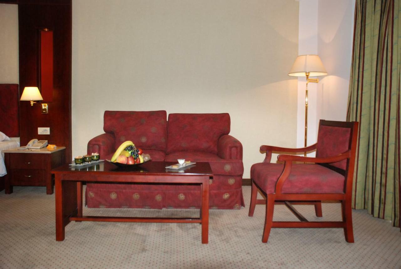 Safir Hotel Cairo Giza Exterior photo The photo shows a cozy seating area in a hotel room. There is a red patterned sofa and a matching armchair placed around a wooden coffee table. On the table, there are some snacks or fruits displayed. A floor lamp is positioned beside the sofa, and t