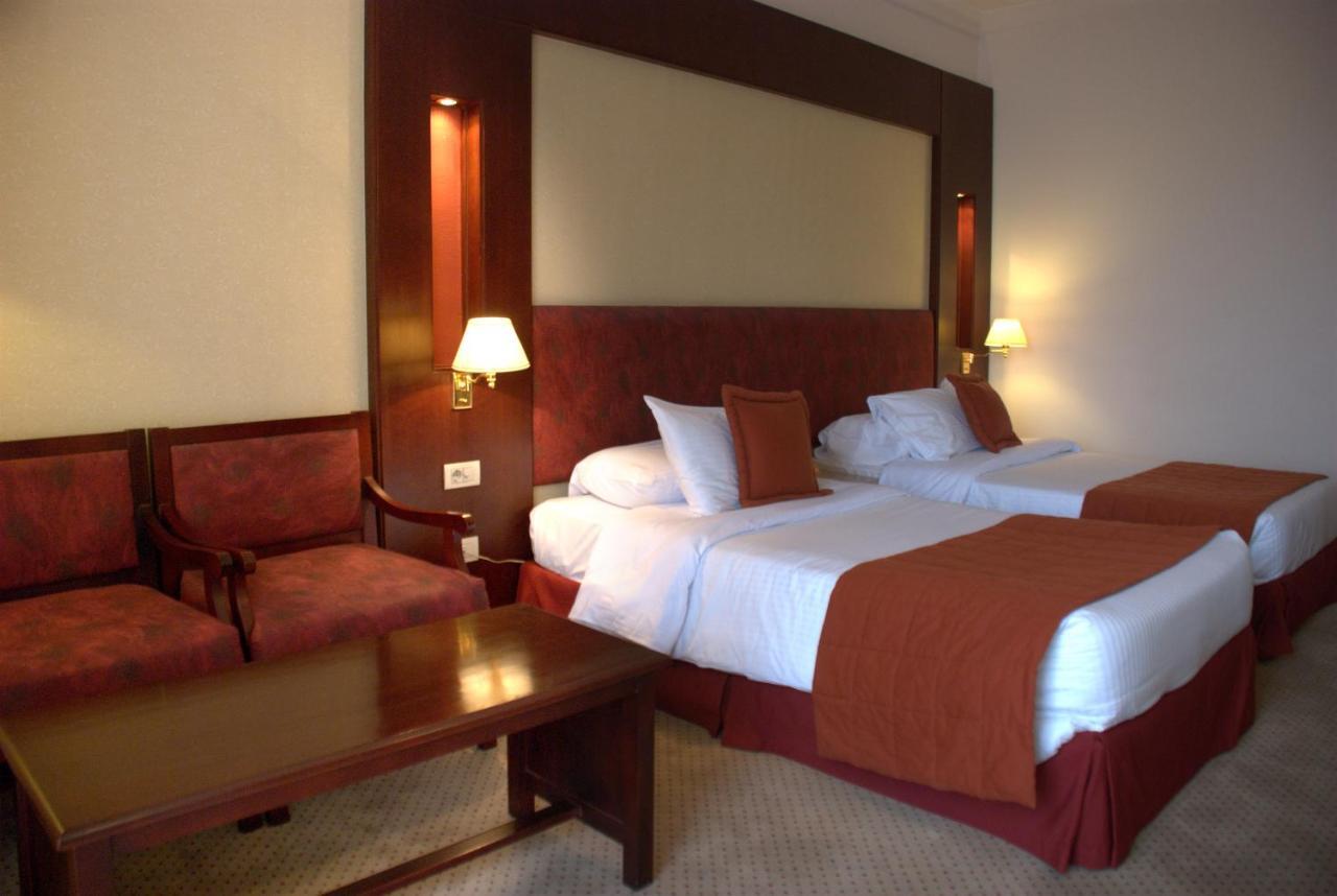 Safir Hotel Cairo Giza Exterior photo The photo shows a hotel room featuring two twin beds with white linens and orange bedspreads. There is a headboard behind the beds, along with wall-mounted lamps on either side. In the corner, there are two upholstered chairs and a wooden coffee tabl